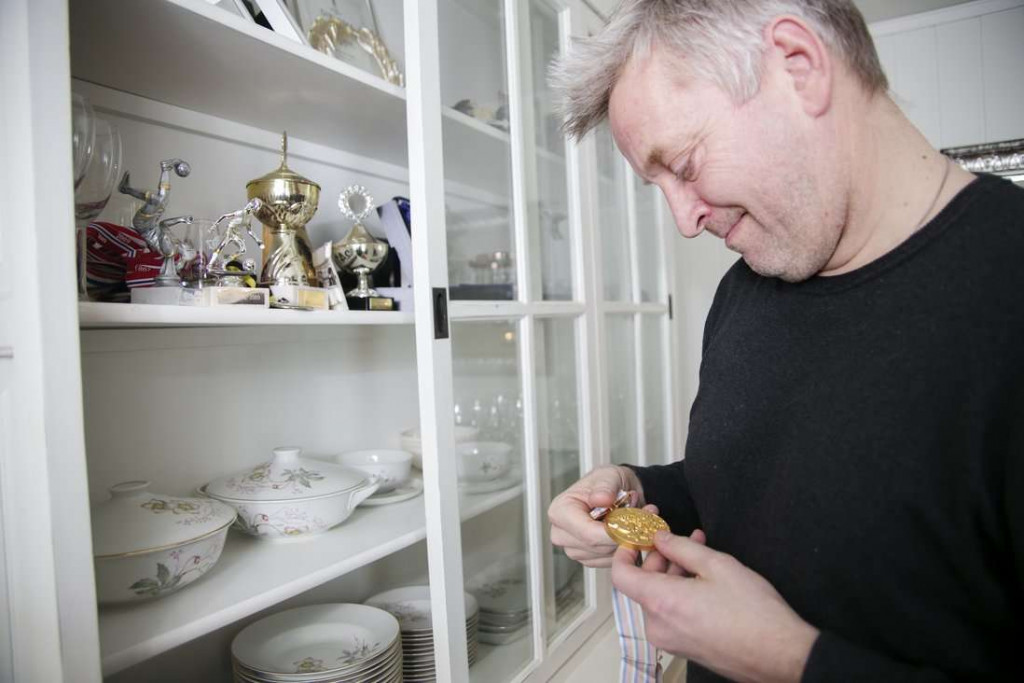 Kjetil står ved siden av et skap fullt av trofeer, og ser på en medalje