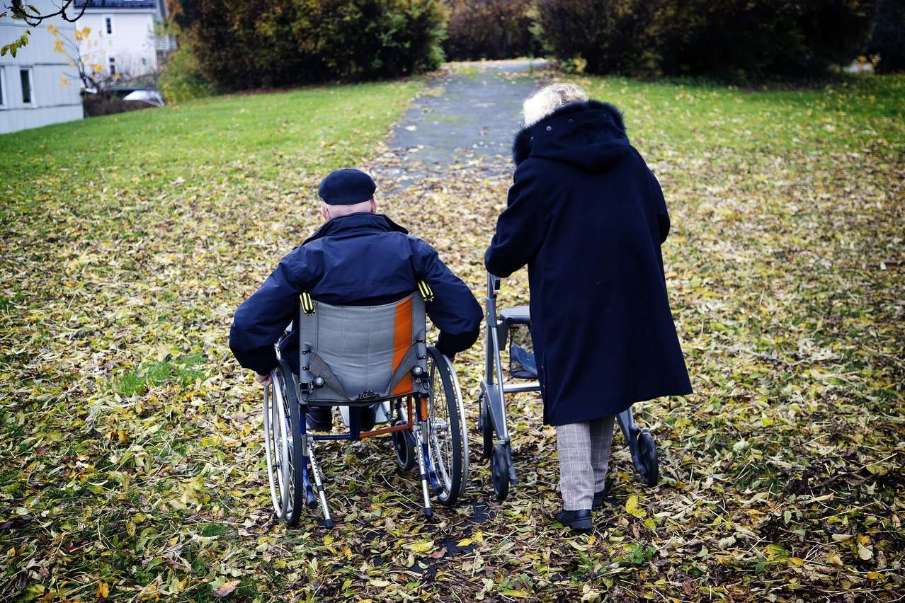 En dame med rullator og en mann i rullestol på tur i parken. Foto.