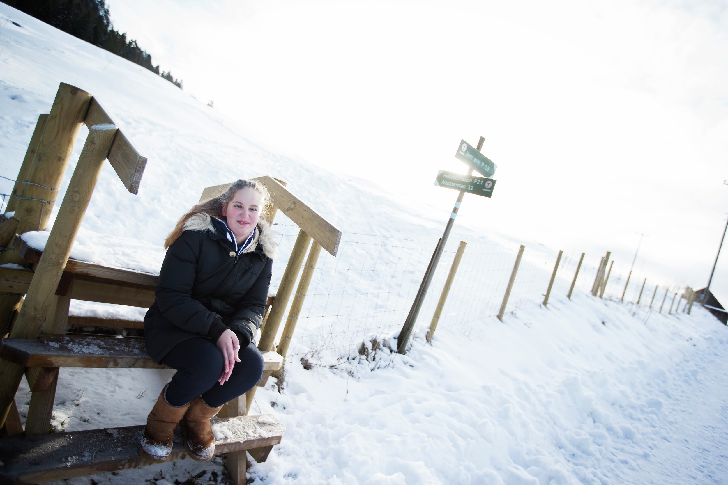 Jente sitter ved gjerde i snøkledd landskap. Foto.