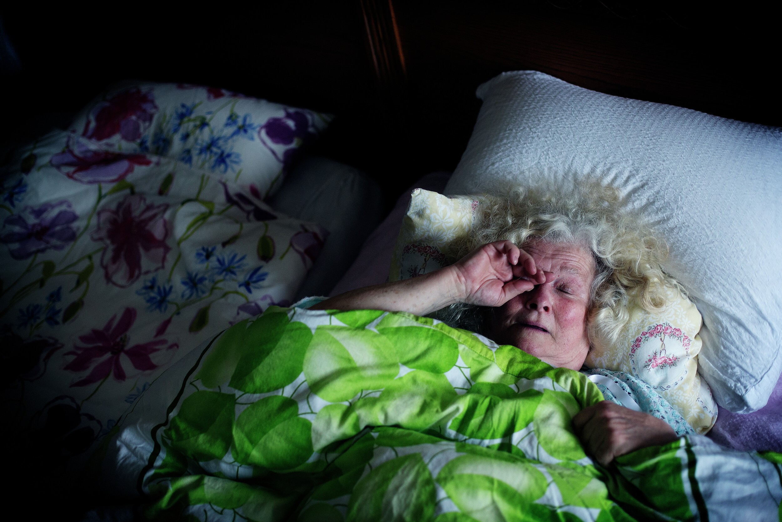 Eldre kvinne ligger i senga og gnir seg i øynene. Foto.