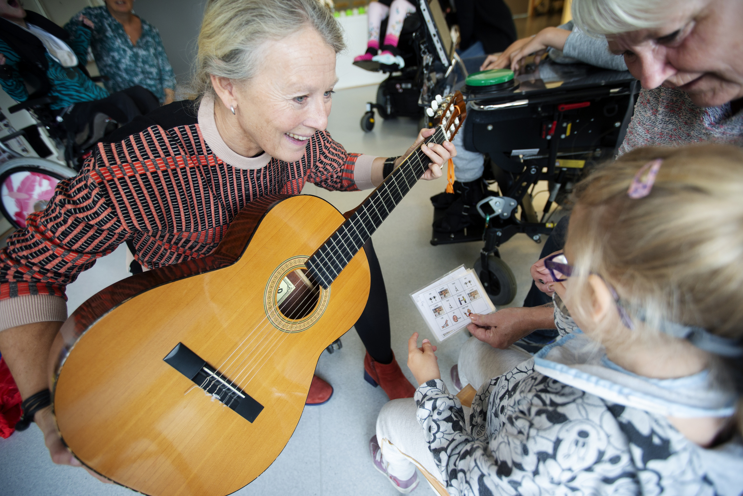 Kvinne med gitar smiler mot barn og eldre kvinne. Foto.