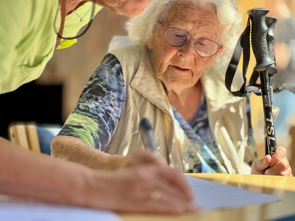 Eldre kvinne med staver i hånd sitter ved bord og skriver. Foto