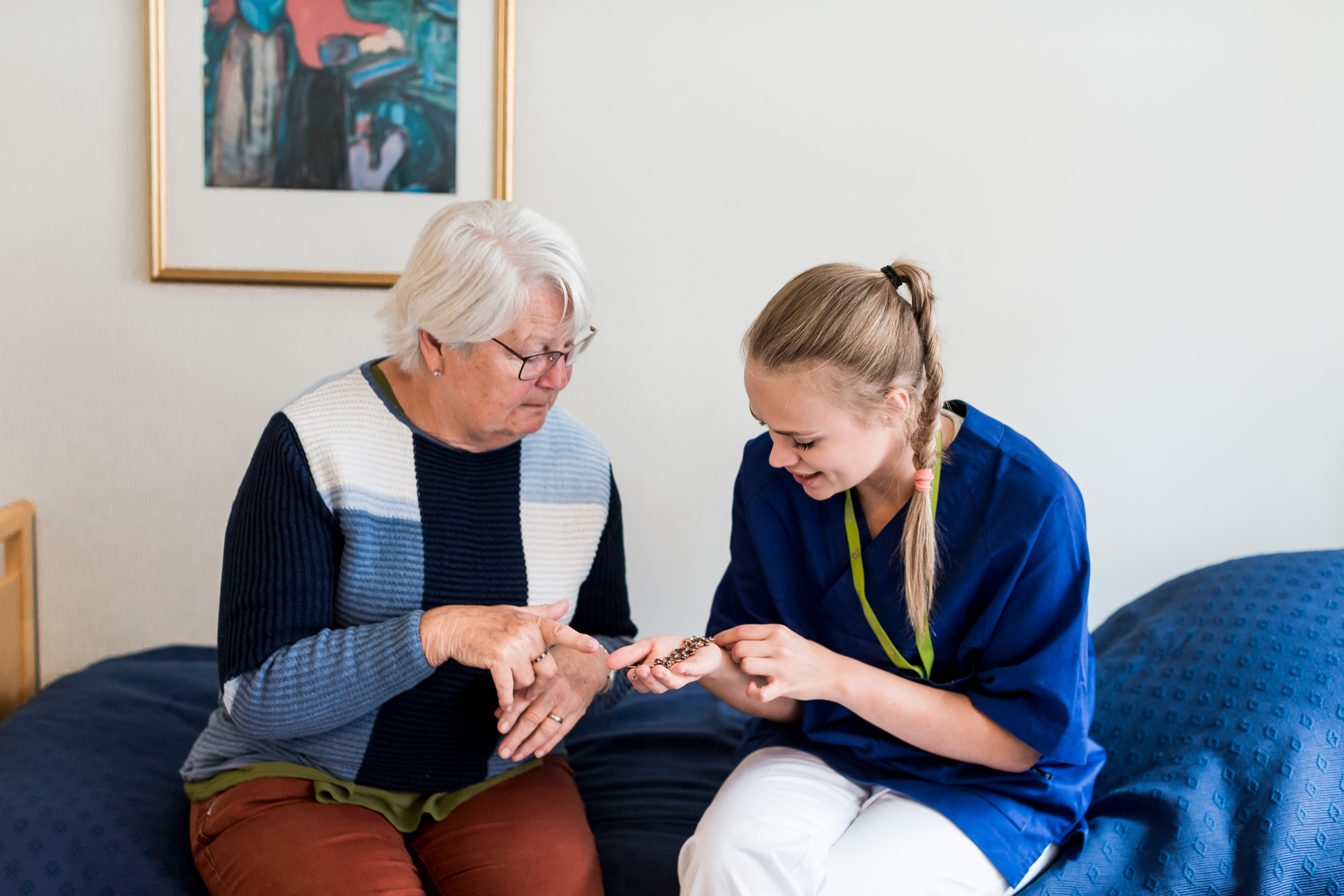 Eldre dame og sykepleier sitter på en seng og ser på en liten gjenstand.