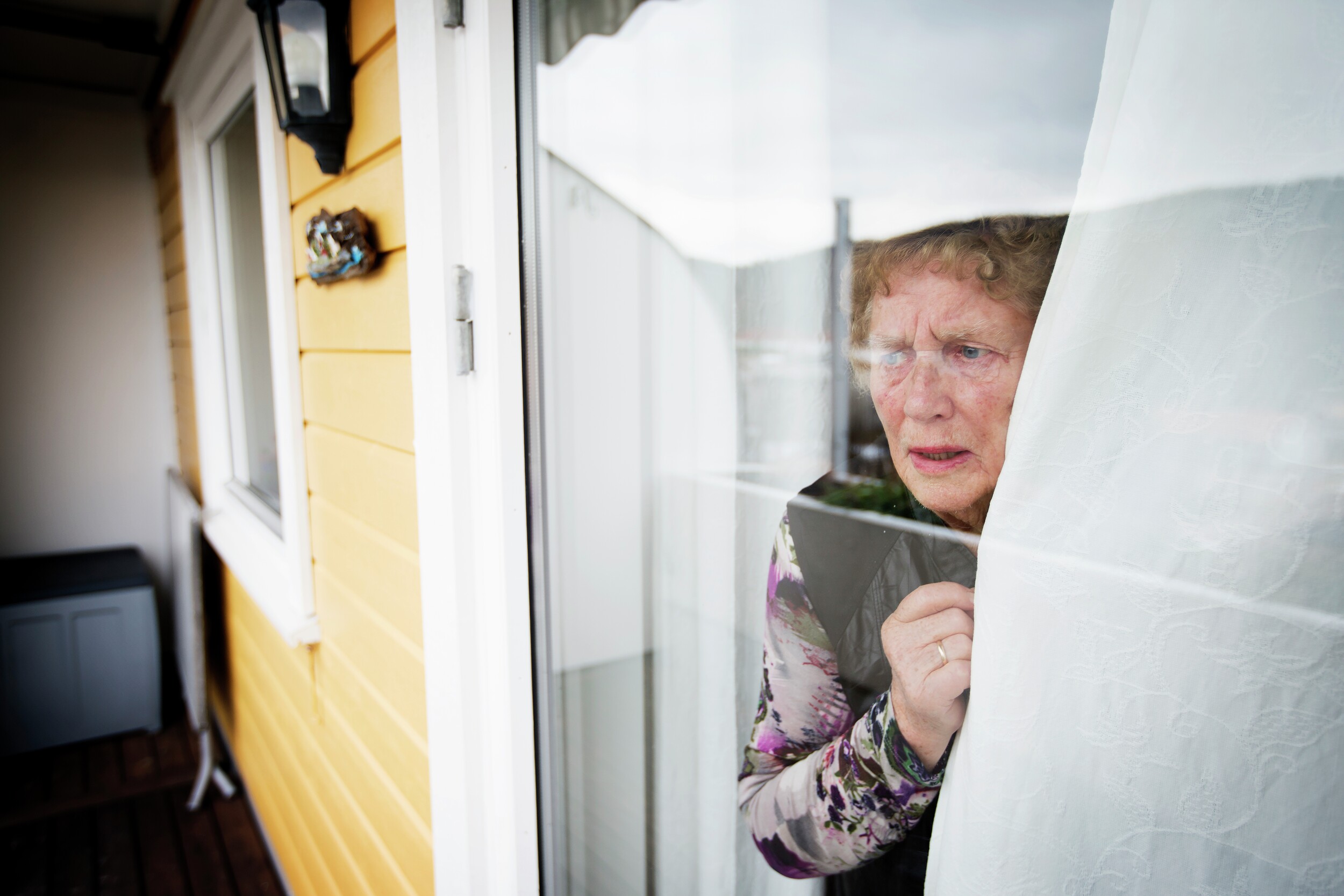 Redd eldre dame står i vinduet sitt. Foto.