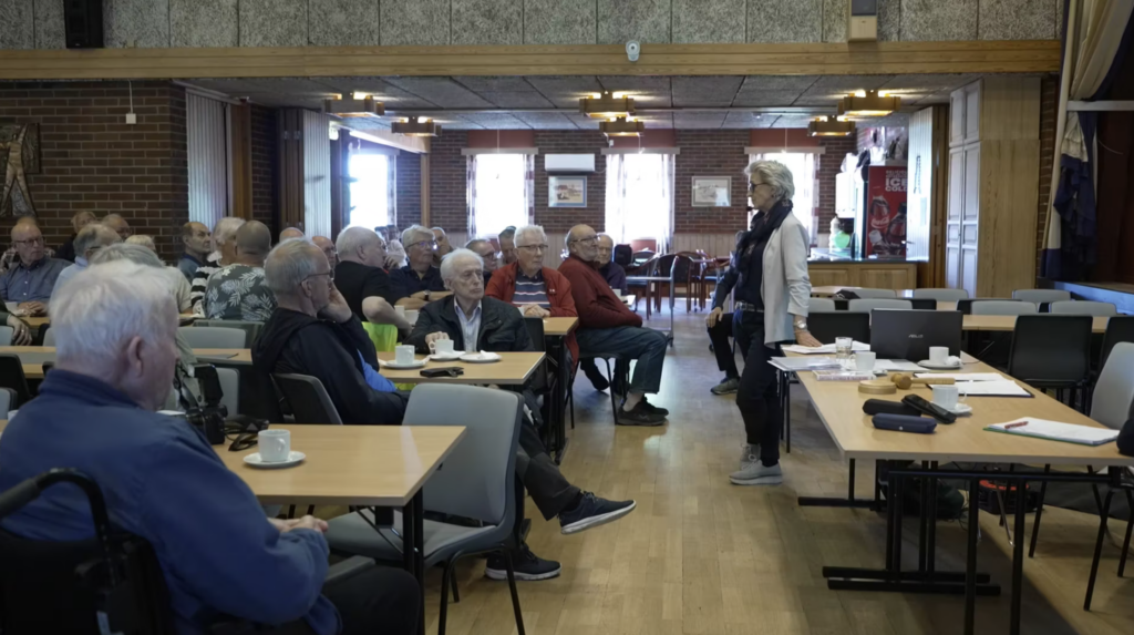 Kvinne snakker til en forsamling eldre menn.